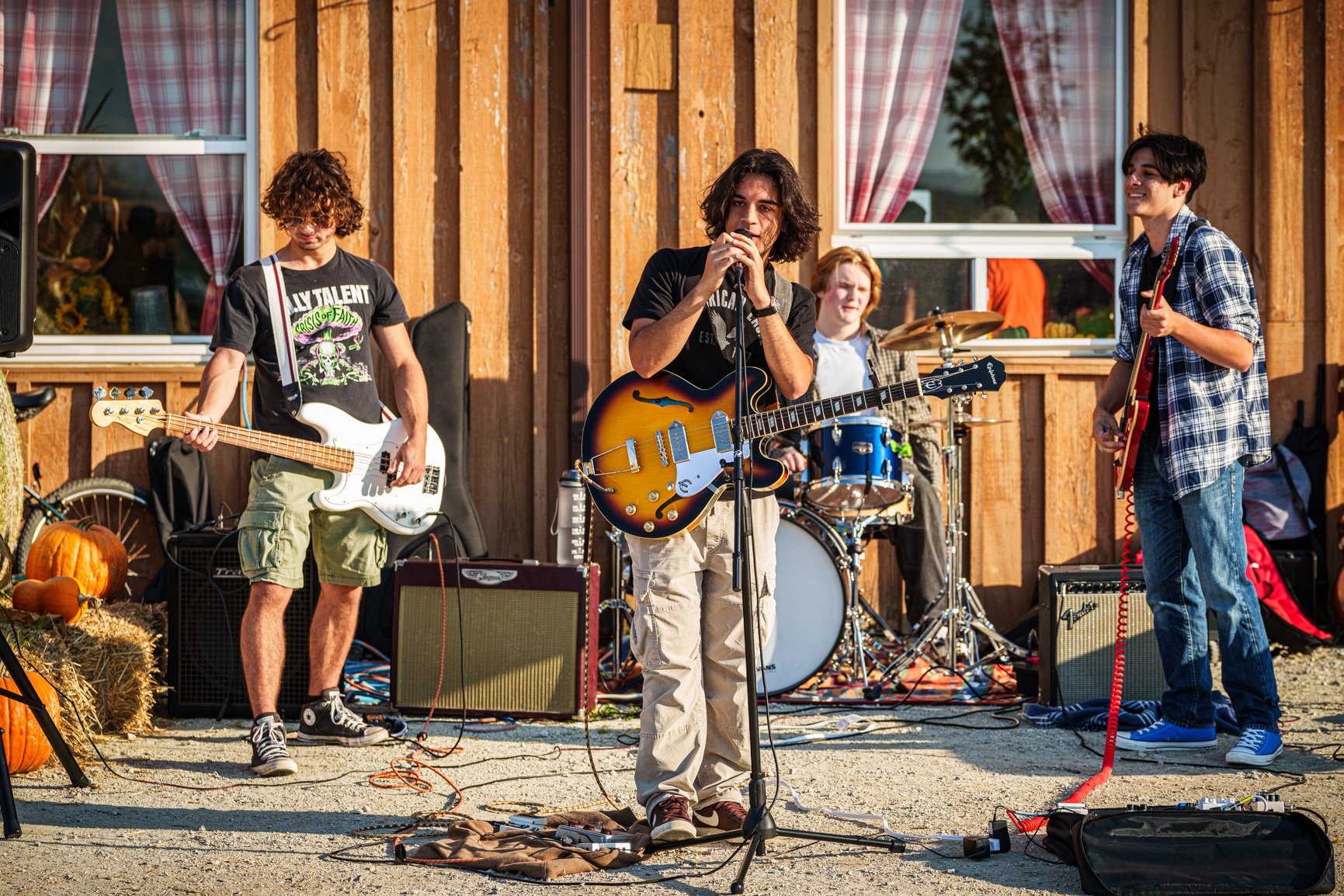 A youth band, performing in frotn of a wooden building, with a three guitarists and one drummer, on a sunny summer day