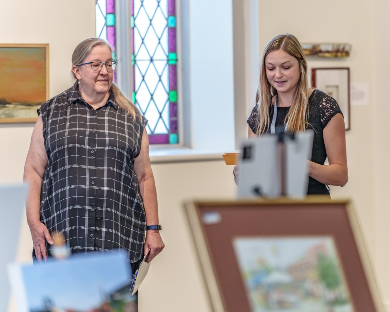 Two adults stand indoors near an easel and drawing setup. One adult looks toward the other, who is reading from a small paper or card; framed artwork and a stained-glass style window are visible in the background.