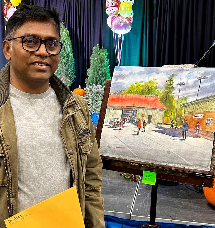 A person wearing glasses, a light shirt, and brown jacket stands indoors beside an easel displaying a first-place winning painting. The painting shows an outdoor scene with buildings, trees, and people. The person holds a yellow envelope, and the background includes colourful balloons, decorations, and greenery.