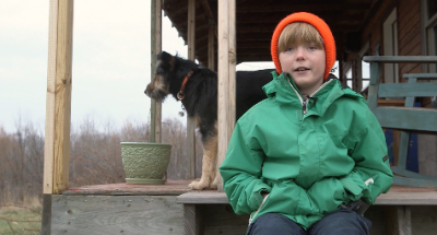 Boy in green jacket sitting on porch with dog behind him