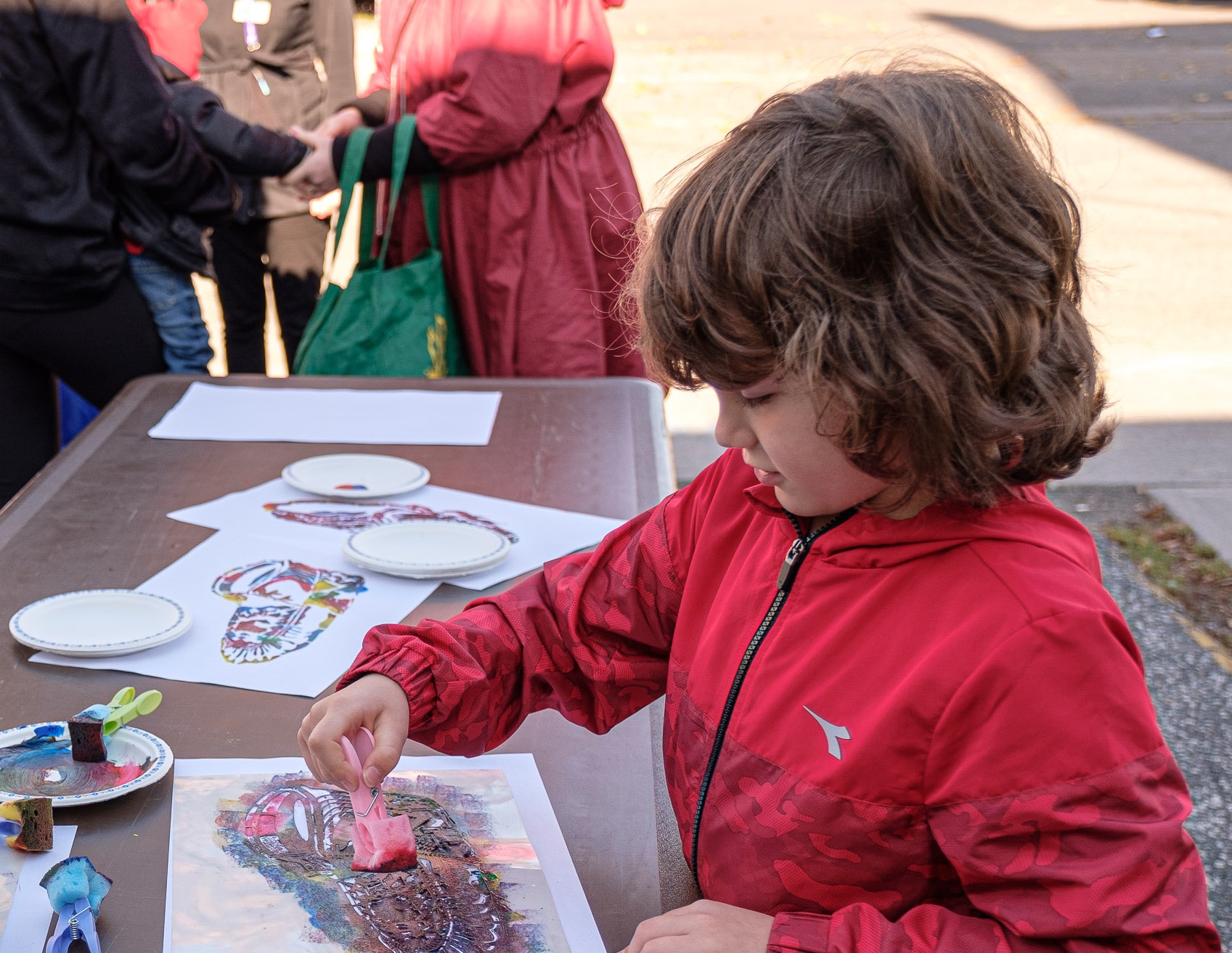 A child wearing a red jacket uses a sponge stamp to apply paint to a printed design on paper at an outdoor activity table. Paint trays, paper plates, and additional printed designs are spread across the table, with other participants and bags visible in the background.