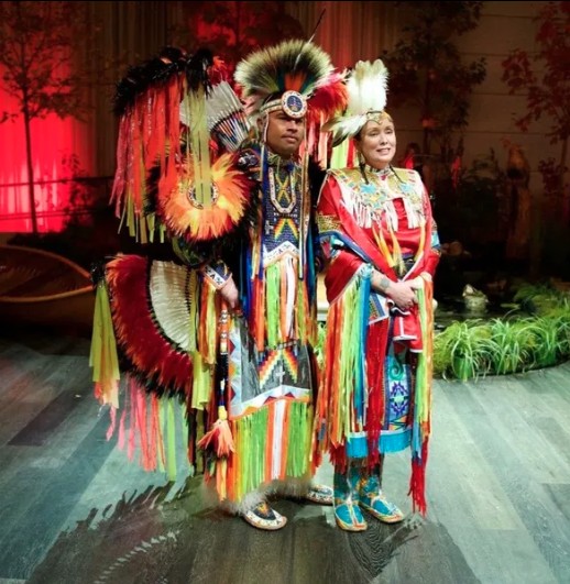 Two performers stand side by side indoors, wearing brightly coloured, elaborately decorated outfits with long flowing fringe, beadwork, and feathered headdresses in red, yellow, green, blue, black, and white. The space is softly lit with red lighting in the background, and plants and wooden flooring are visible around them.