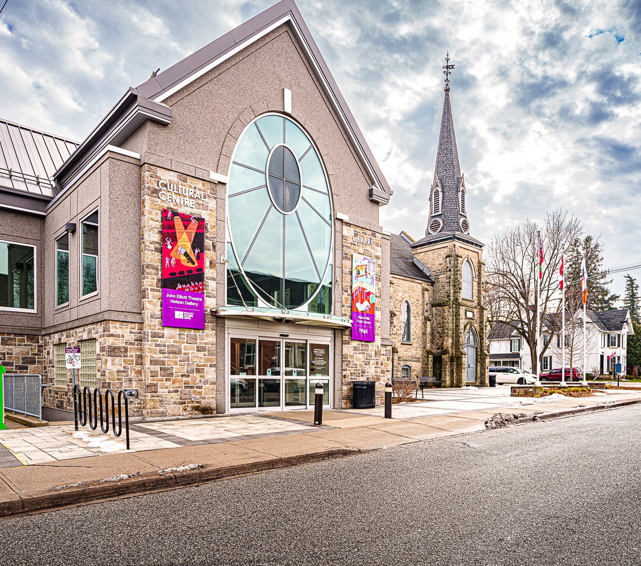 A street‑level view of the library and cultural centre in Halton Hills with stone and grey exterior walls and a large oval window above glass entrance doors. Two colourful vertical banners hang on either side of the entrance. A historic stone church with a tall steeple stands directly beside the building. Several flagpoles with flags are visible in the plaza area to the right, along with nearby houses and bare trees under a cloudy sky